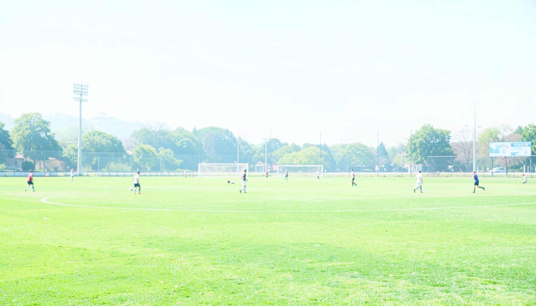 A wide-angle shot of a green football pitch under bright afternoon sunlight, with players warming up in the distance