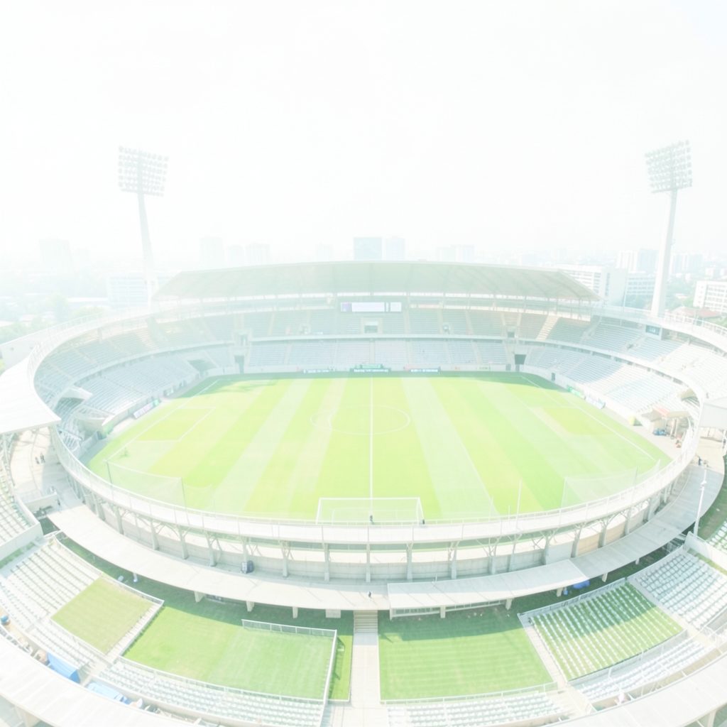 A bright, airy photograph of a municipal football stadium with green pitch and modest stands under a clear sky